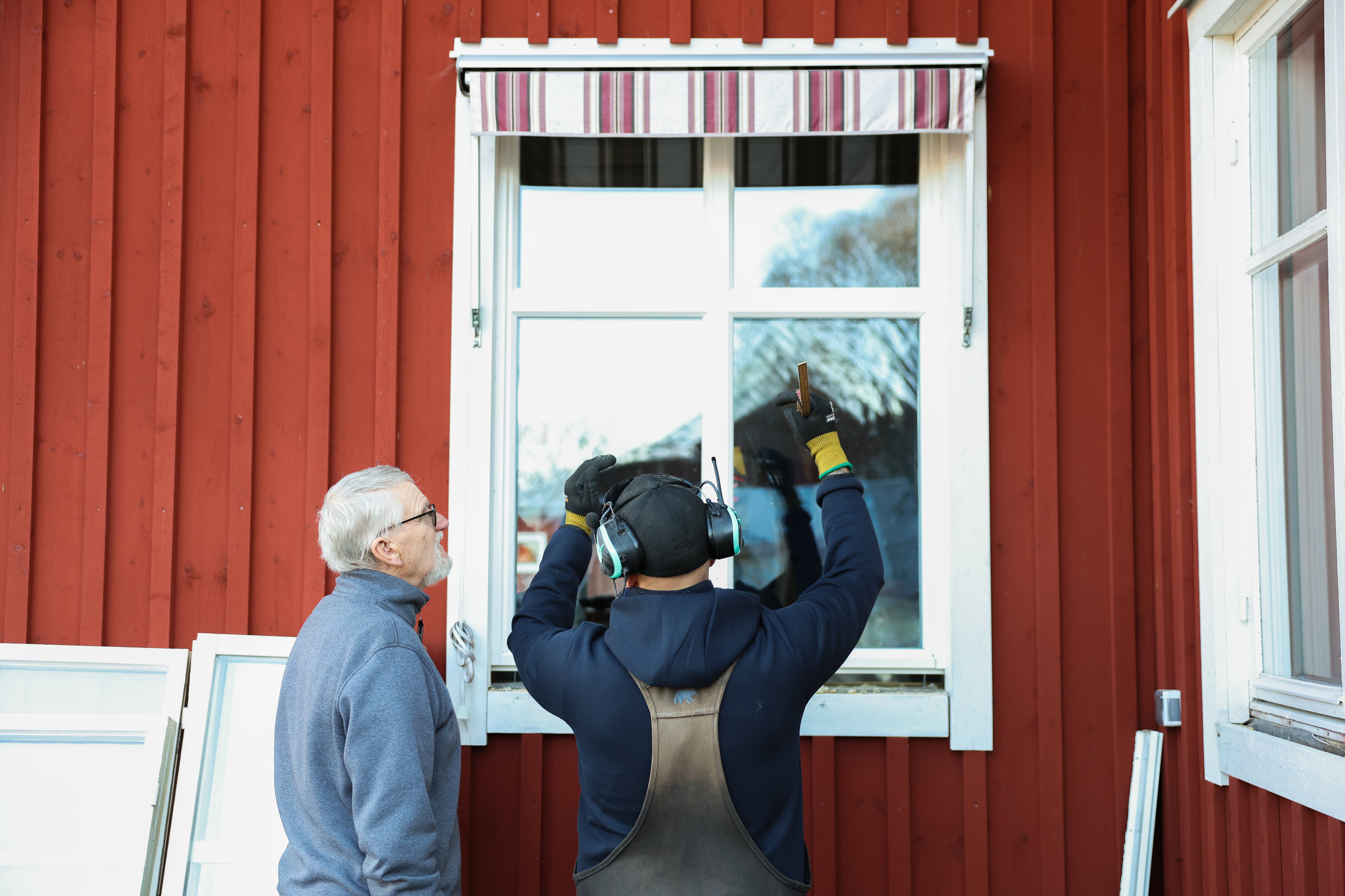 En man monterar ett fönster i ett rött hus och en äldre man står bredvid och tittar på