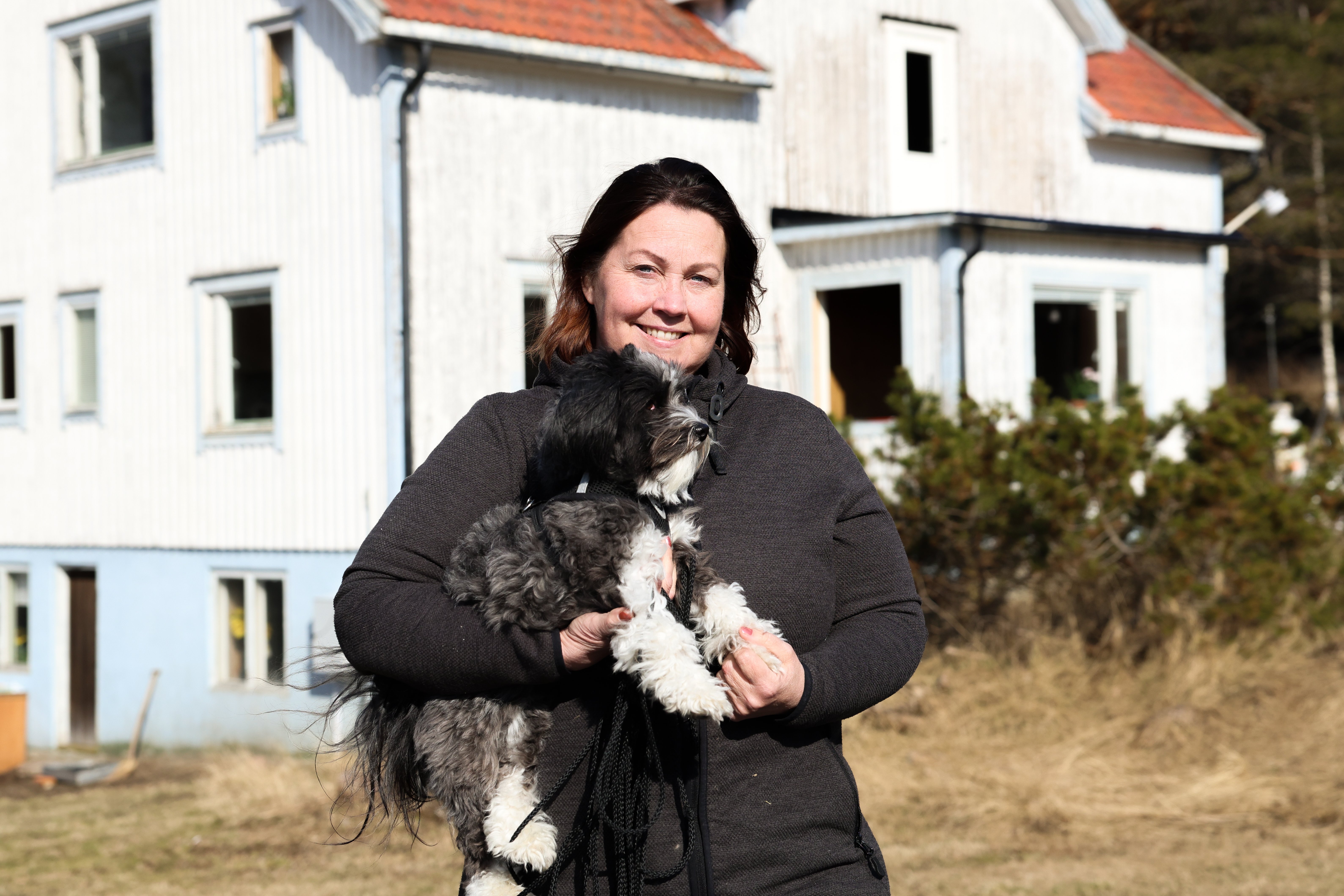Kvinna står med en hund i famnen framför ett vitt hus