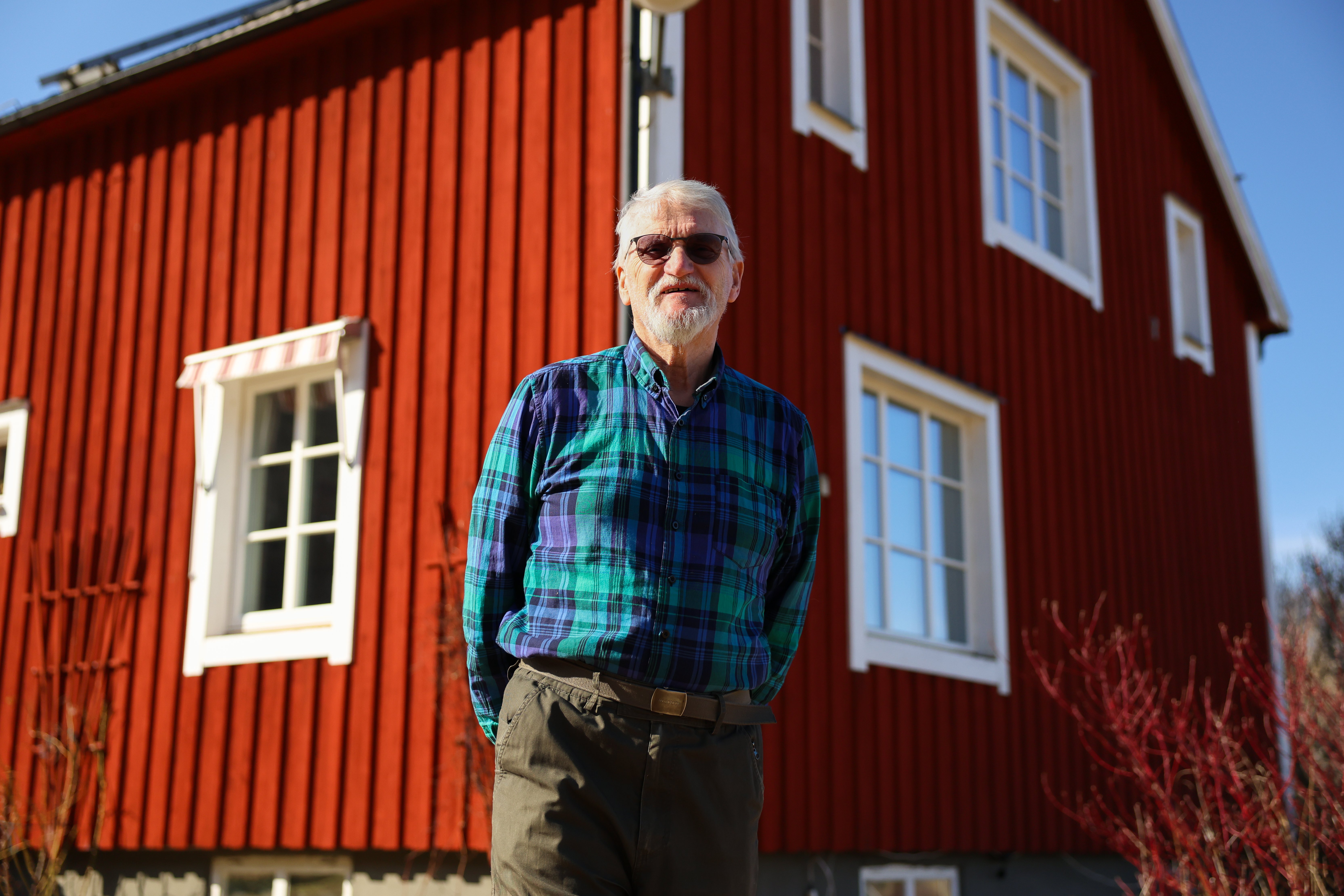 Äldre man i blå rutig skjorta framför ett rött hus med vita knutar