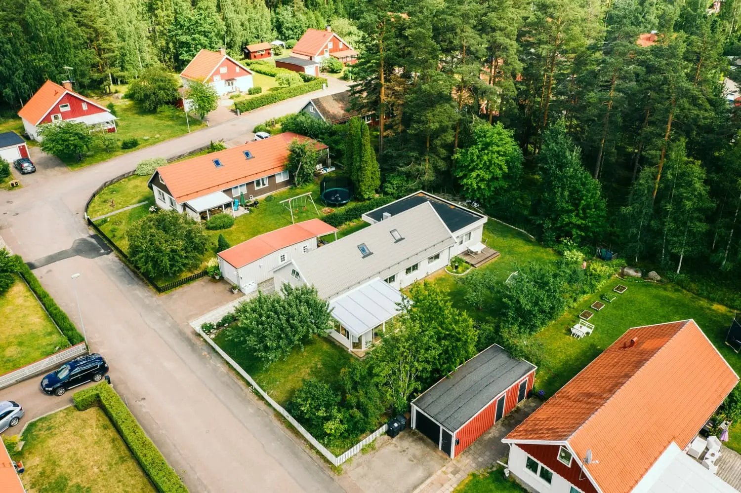 Drönarbild över villakvarter i Falun sommartid med Fias ljusgråa 60-talsvilla i mitten, omgiven av grönskande trädgårdar och tallskog.