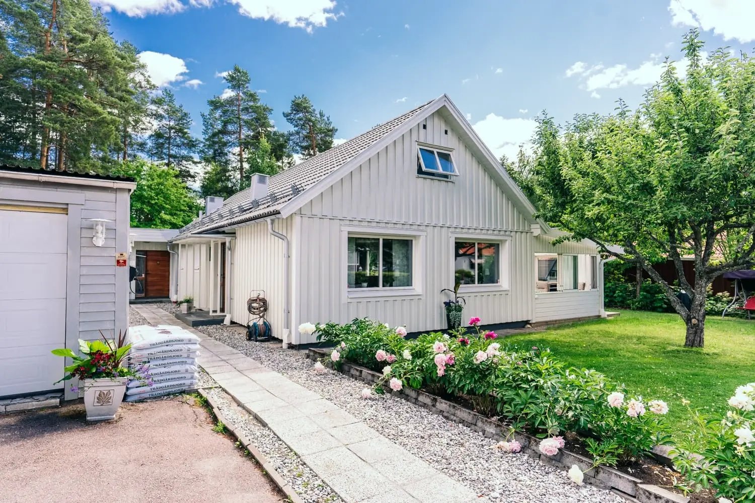 Framsida av renoverad 60-talsvilla i Falun med ljusgrå träpanel, vita energieffektiva fönster från Mockfjärds och blommande pionrabatt längs gångvägen.