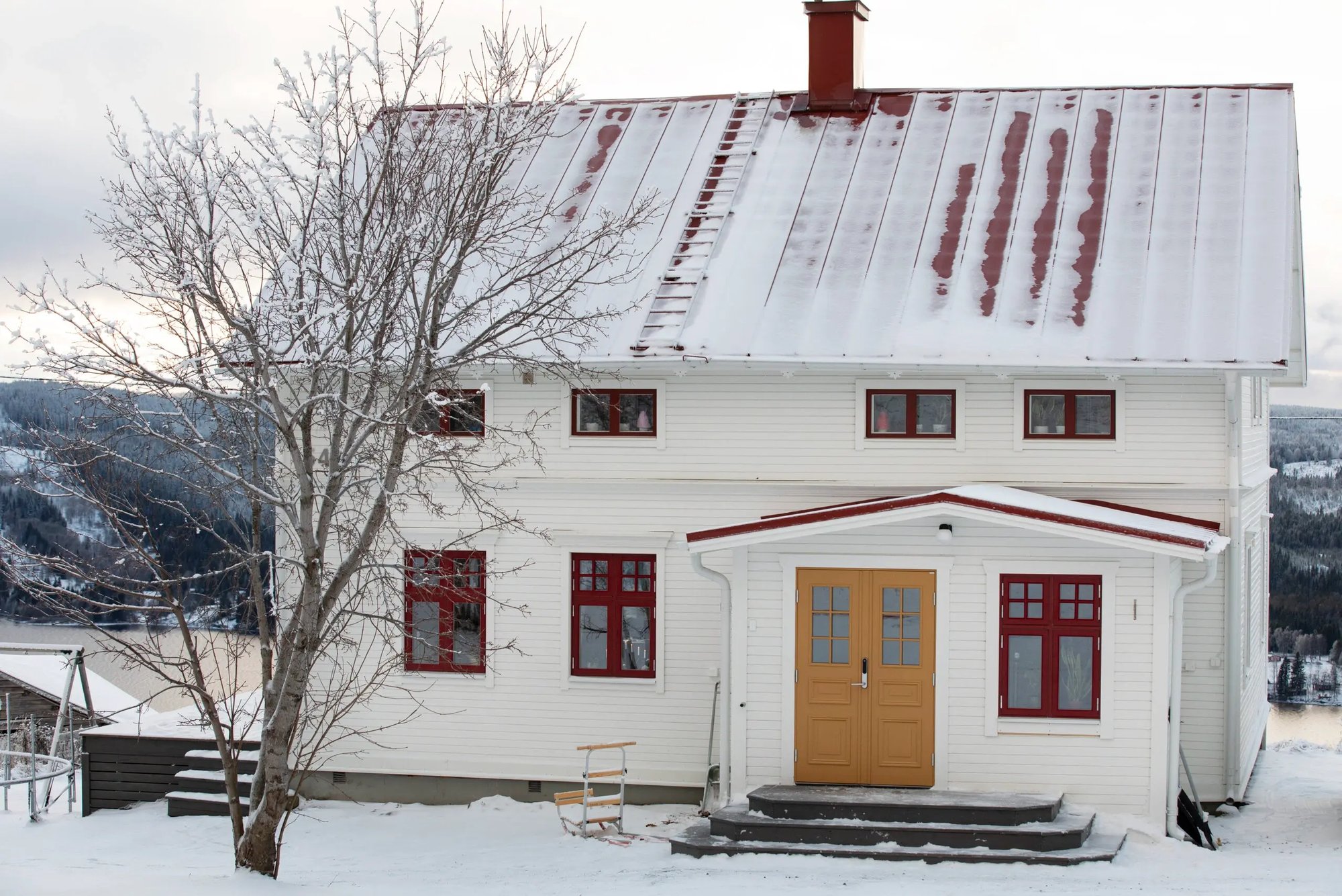 Vitt trähus från 1901 i vinterlandskap med snötäckt tak, röda fönster och ockrafärgad pardörr, beläget i Kaxås Offerdal i Jämtland.
