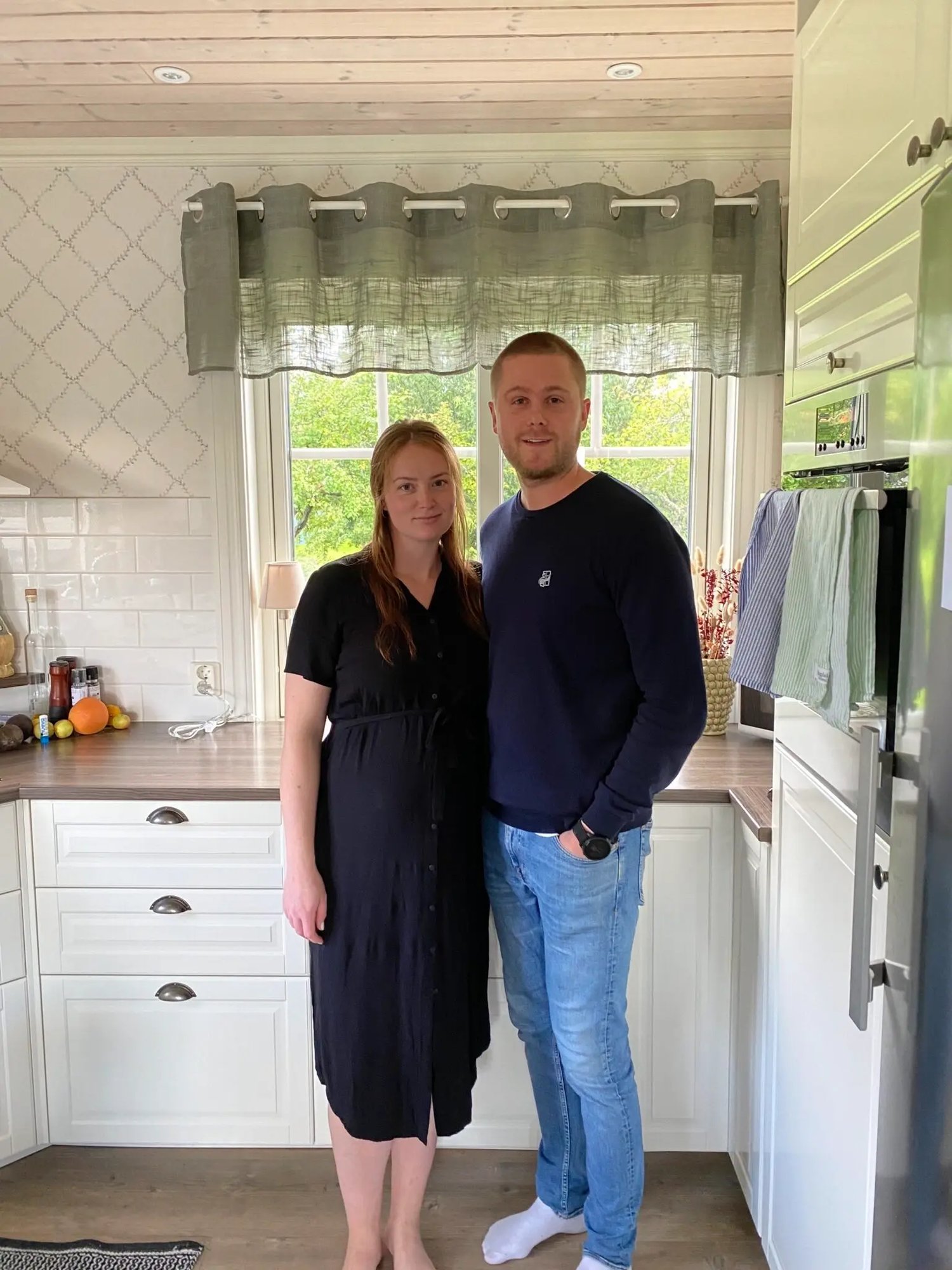 Erik och Emma står tillsammans i sitt renoverade kök i Leksand framför ett nytt vitt vridfönster från Mockfjärds med utsikt mot trädgården.