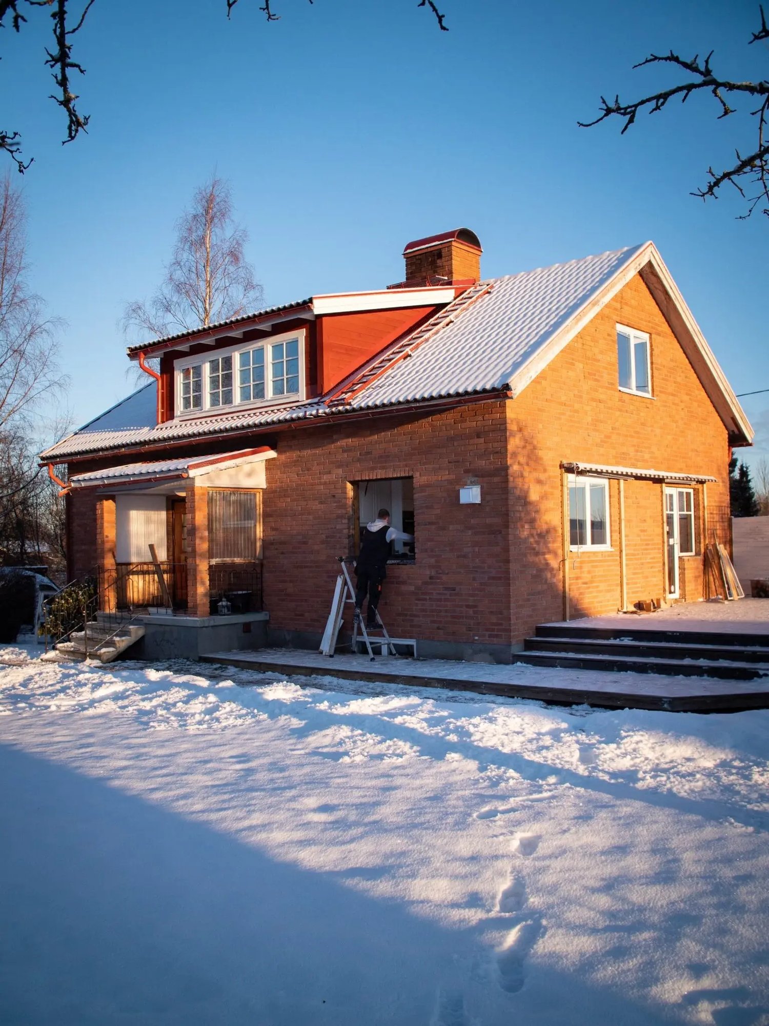 Rött tegelhus från 1956 i Leksand under pågående fönsterbyte i januari med montör på stege, snötäckt tomt och klart vinterhimmel.