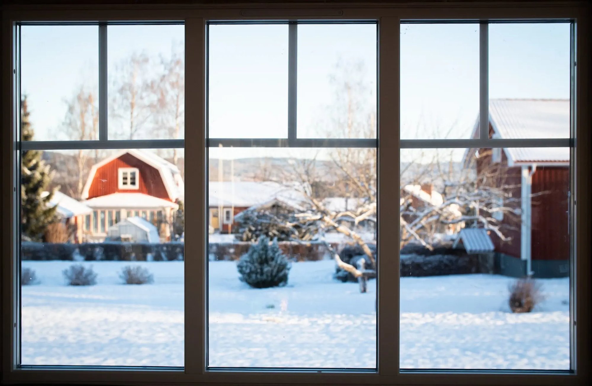 Utsikt genom ett nytt fast köks­fönster med tre stående poster från Mockfjärds i Eriks och Emmas tegelhus, med snötäckt trädgård och röda hus i Leksand utanför.