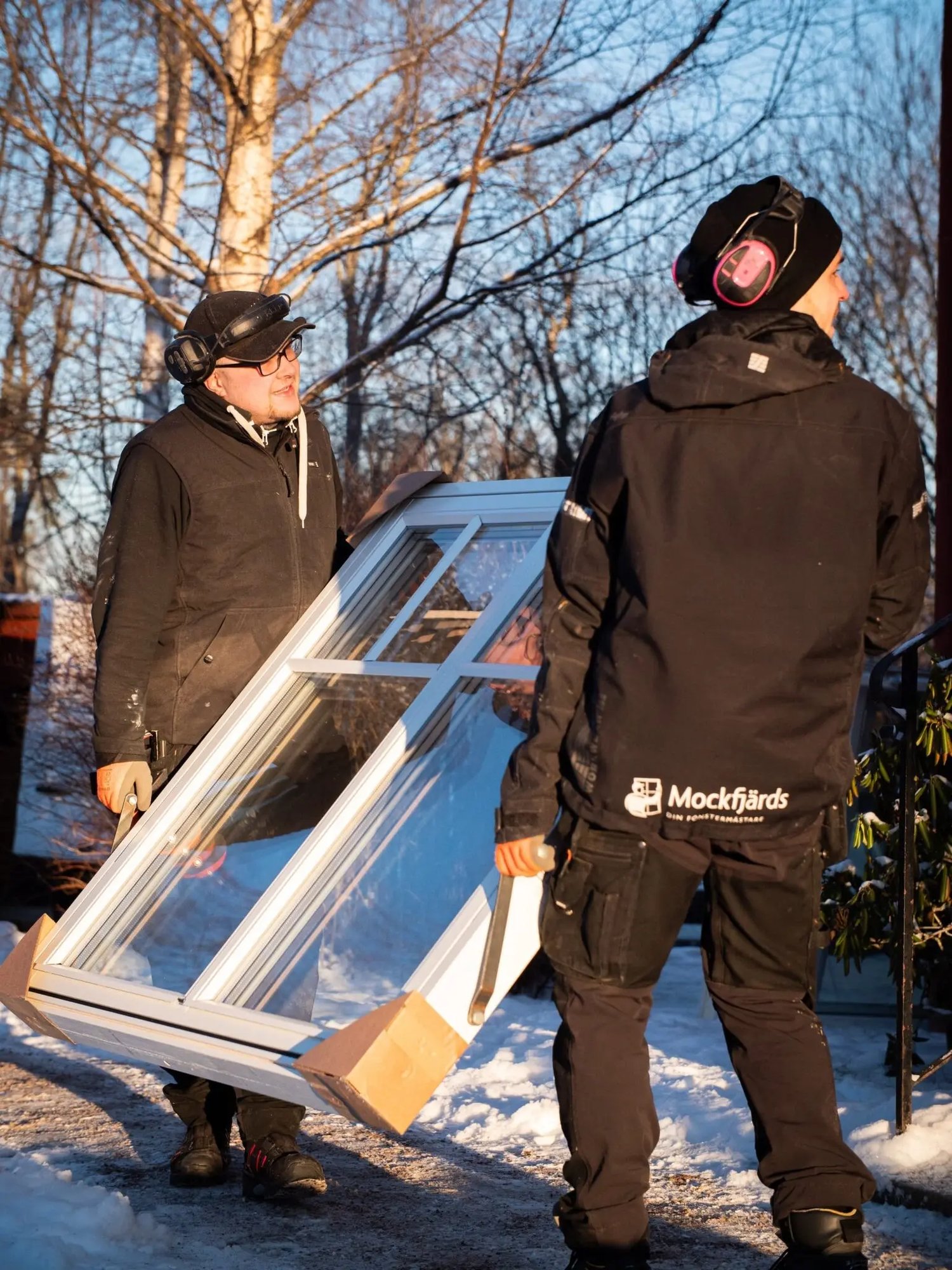 Två Mockfjärds-montörer bär ett nytt vitt fönster med spröjs i vintermiljö utanför tegelhuset i Leksand under fönsterbytet i januari.