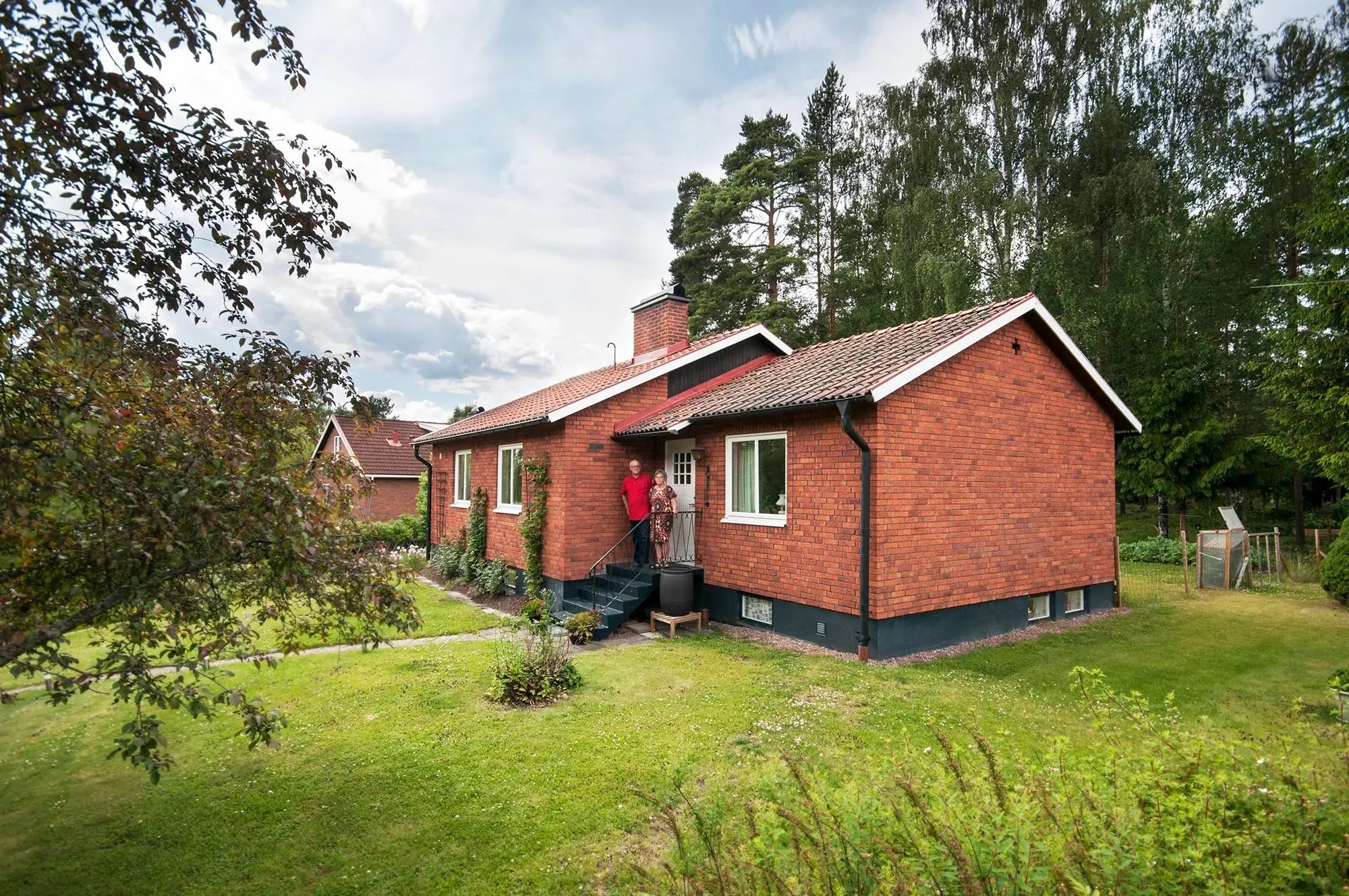 Åke Pers och Ann-Mari Nilsson står vid entrén till sin röda tegelvilla från 1960 i Hosjö utanför Falun, med nya vita Prima-fönster från Mockfjärds och frodig sommargrönskan trädgård.