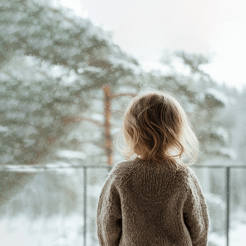 Litet barn står vid ett fönster och tittar ut på fallande snö.
