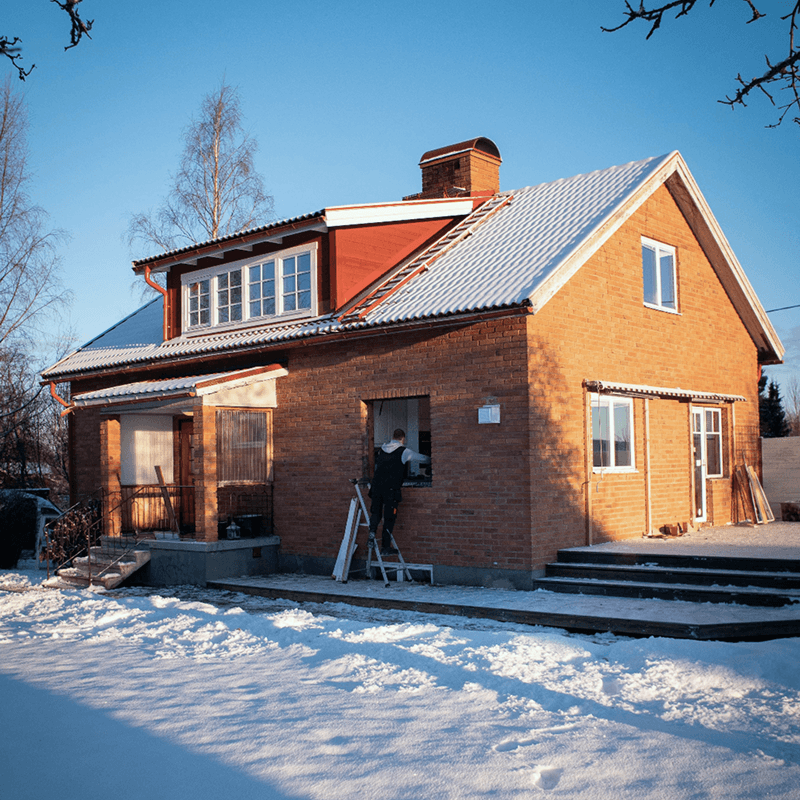 Person står på en stege och installerar ett fönster i ett tegelhus med snö på marken.