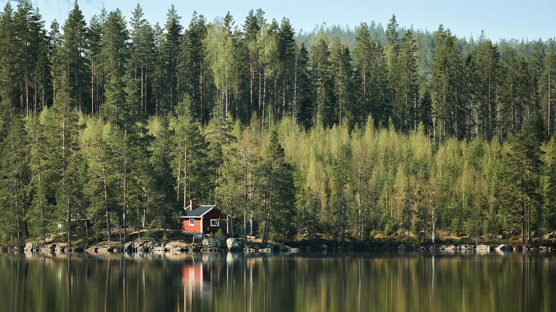 Liten röd stuga vid en sjö omgiven av skog, med spegling i vattnet.