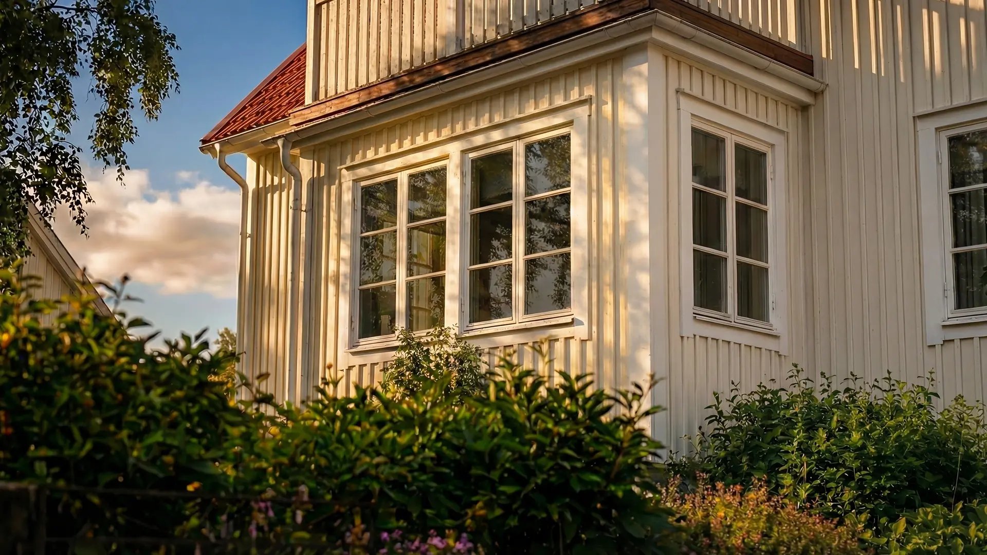 Flera fönster på ett ljust trähus omgivna av grönska i kvällsljus.