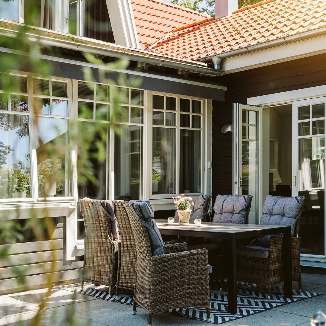 En uteplats med matbord och korgstolar intill ett hus med stora fönster och ett rött tegeltak, med en öppen glasdörr som leder in i huset.
