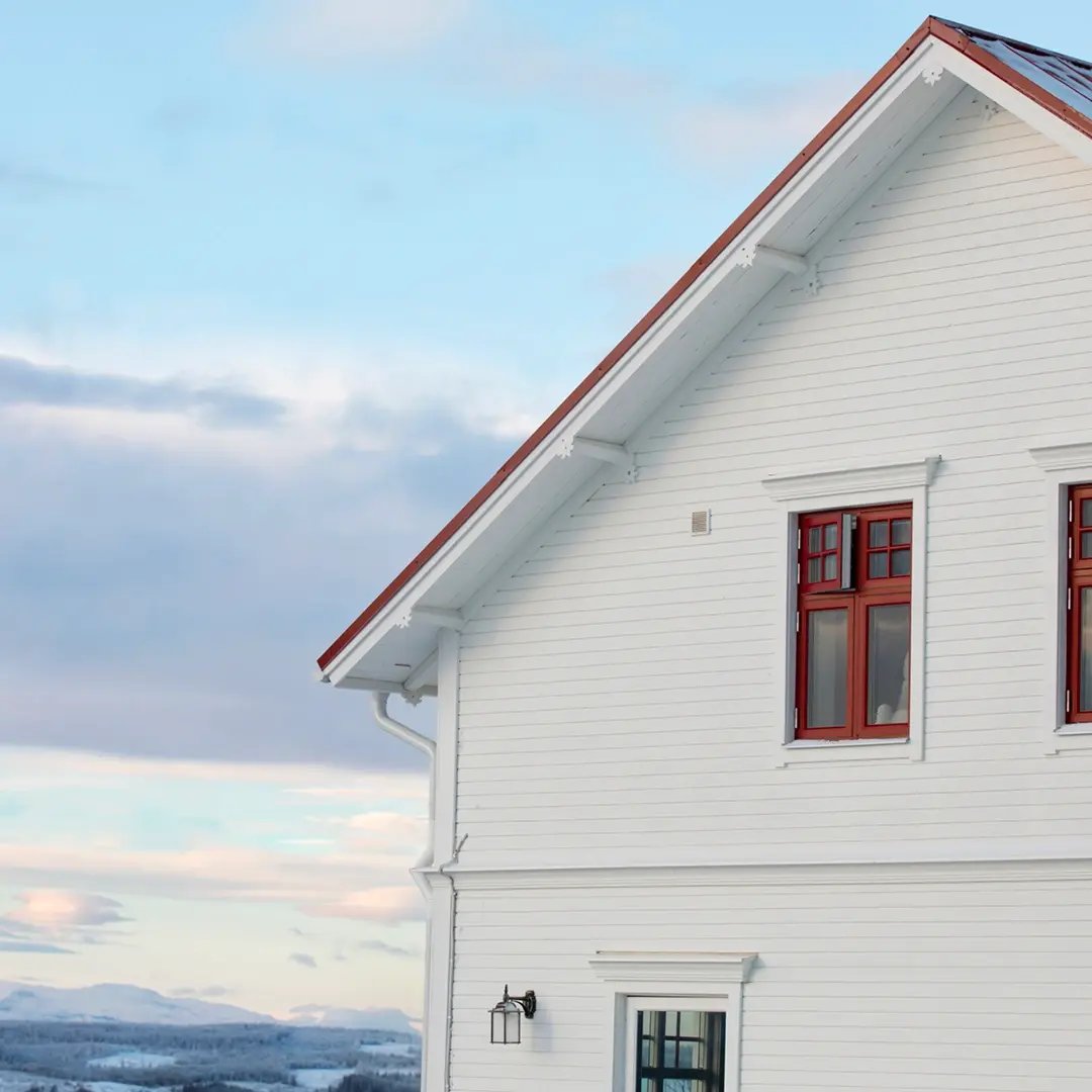 Vit träfasad på ett hus med röda fönster och utsikt över ett vinterlandskap.