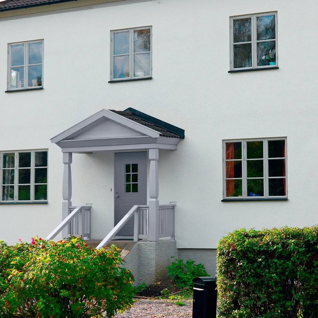 Framsidan av ett vitt tvåvåningshus med en liten täckt veranda, grå ytterdörr, symmetriska fönster och gröna buskar i trädgården.