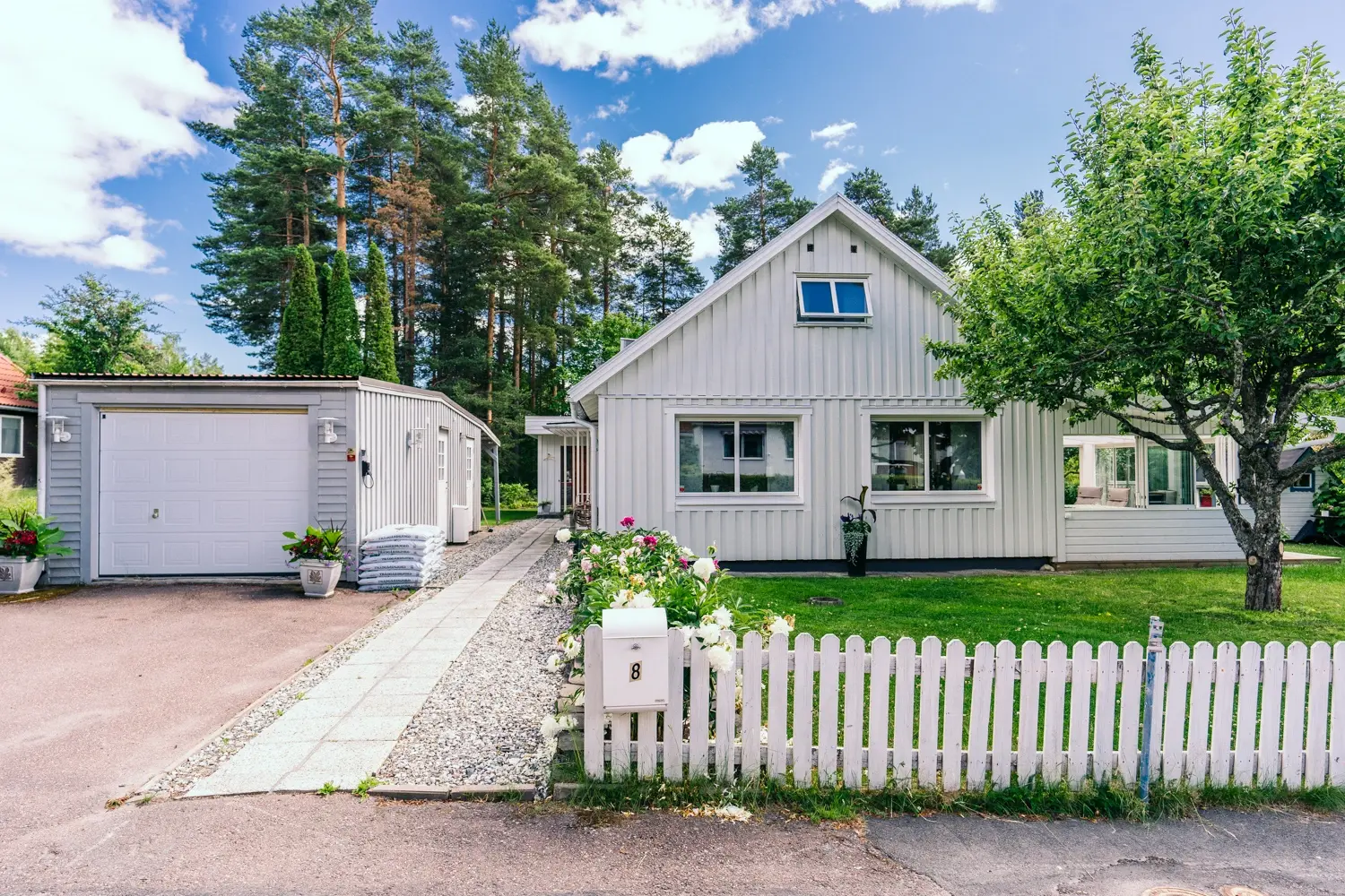  Ljusgrå 60-talsvilla i Falun med ny vit träpanel, vita Mockfjärdsfönster och vit staketinhägnad trädgård med blommande pioner vid ingången.