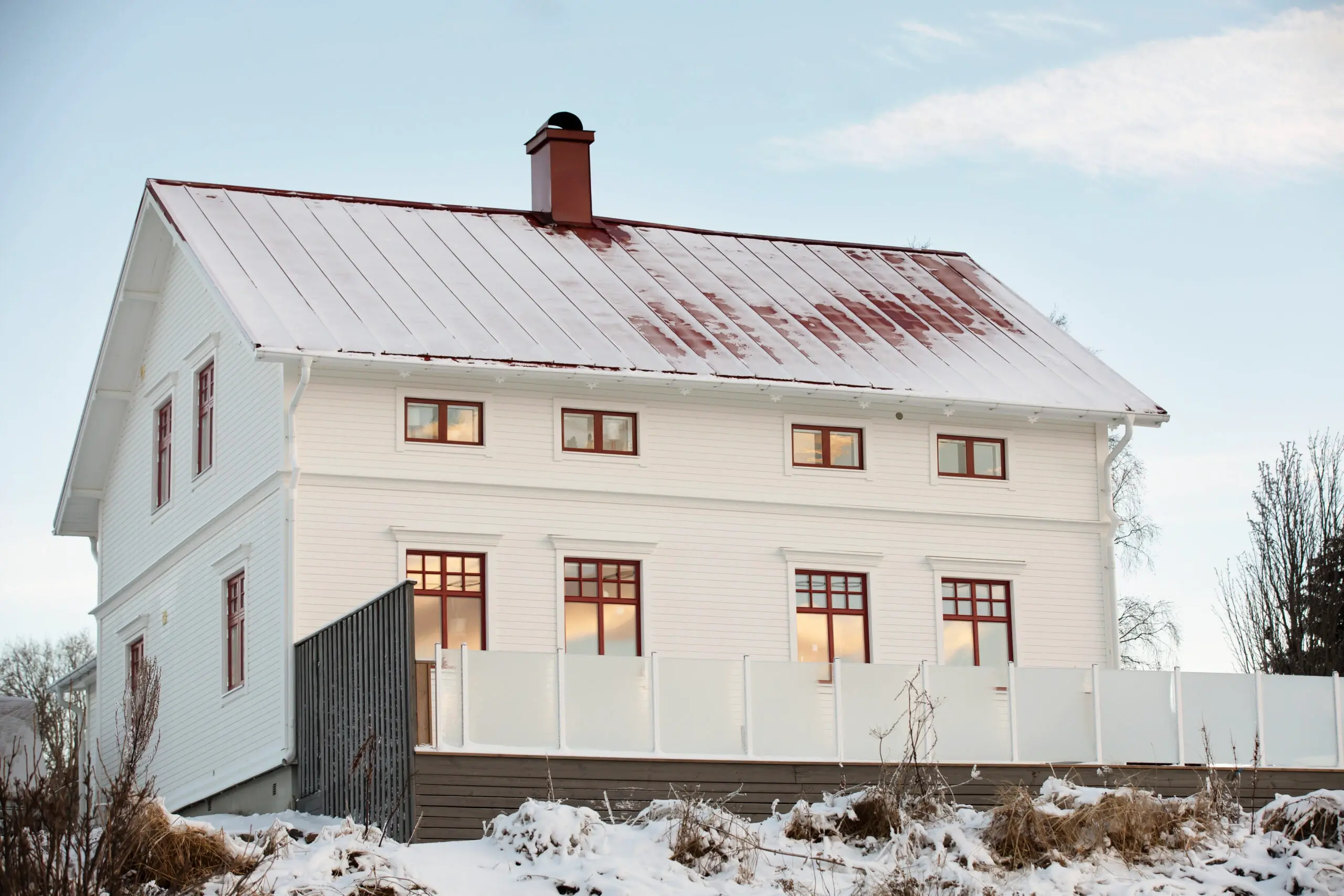 Baksida av vitt sekelskifteshus med röda fönster från Mockfjärds i vintermiljö, med utsikt mot öppet landskap och lätt snötäckt mark.