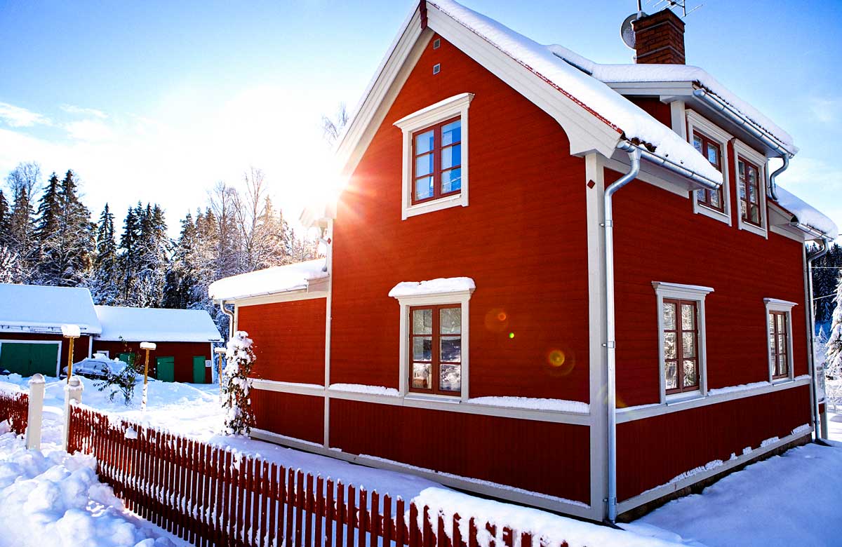 Rött trähus i vintermiljö med snö och solljus.