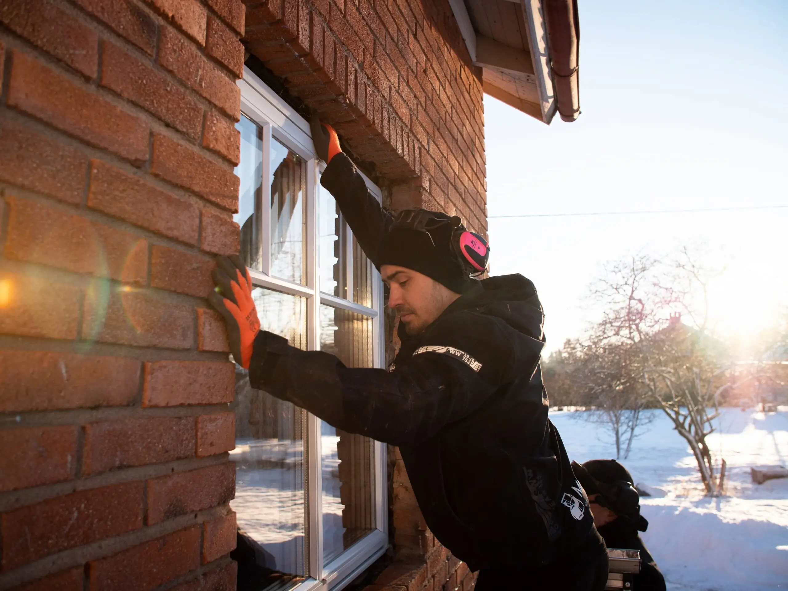 Mockfjärds-montör med orange handskar monterar ett nytt vitt fönster utifrån i tegelfasaden på Eriks och Emmas hus i Leksand vid minusgrader.