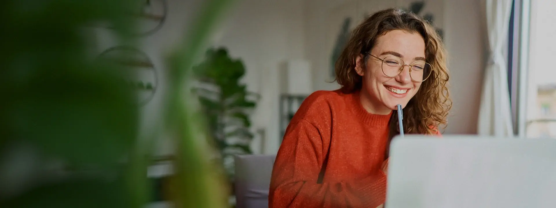 Kvinna sitter hemma vid dator och planerar, ler medan hon arbetar i en ljus inomhusmiljö.