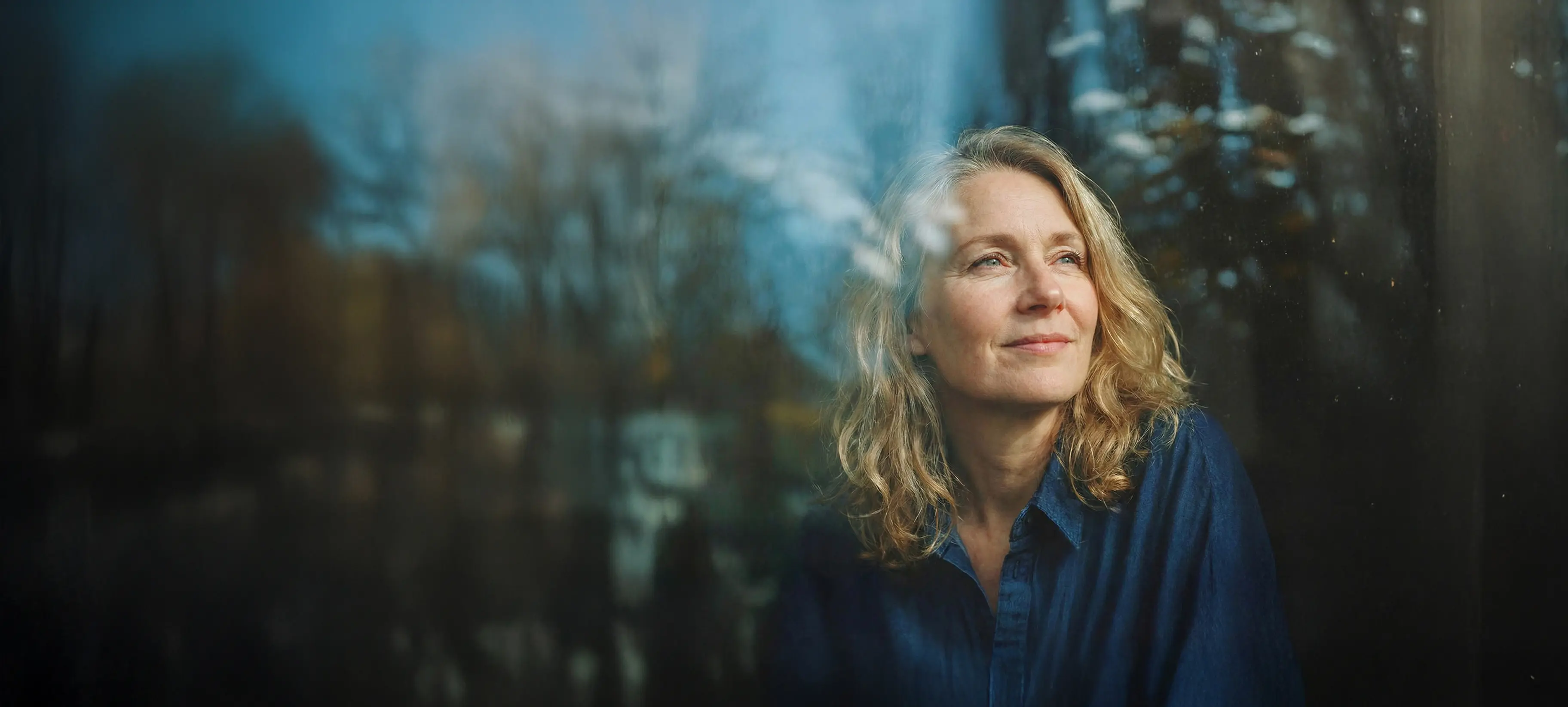 Kvinna sitter vid ett fönster och ser ut, med träd reflekterade i glaset.