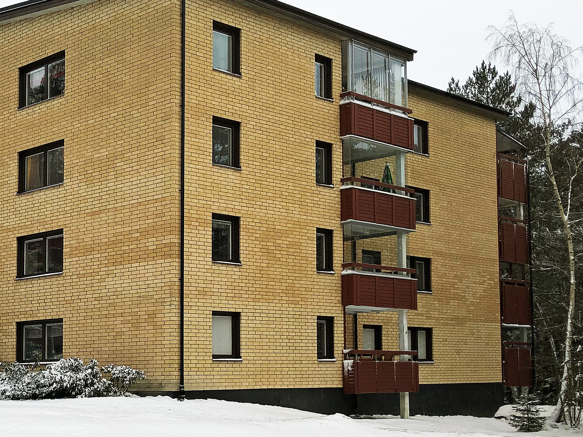 Flervåningshus med gul tegelfasad, röda balkonger och inglasade toppbalkonger i vintermiljö – byte av ca 900 fönster utfört av Mockfjärds.