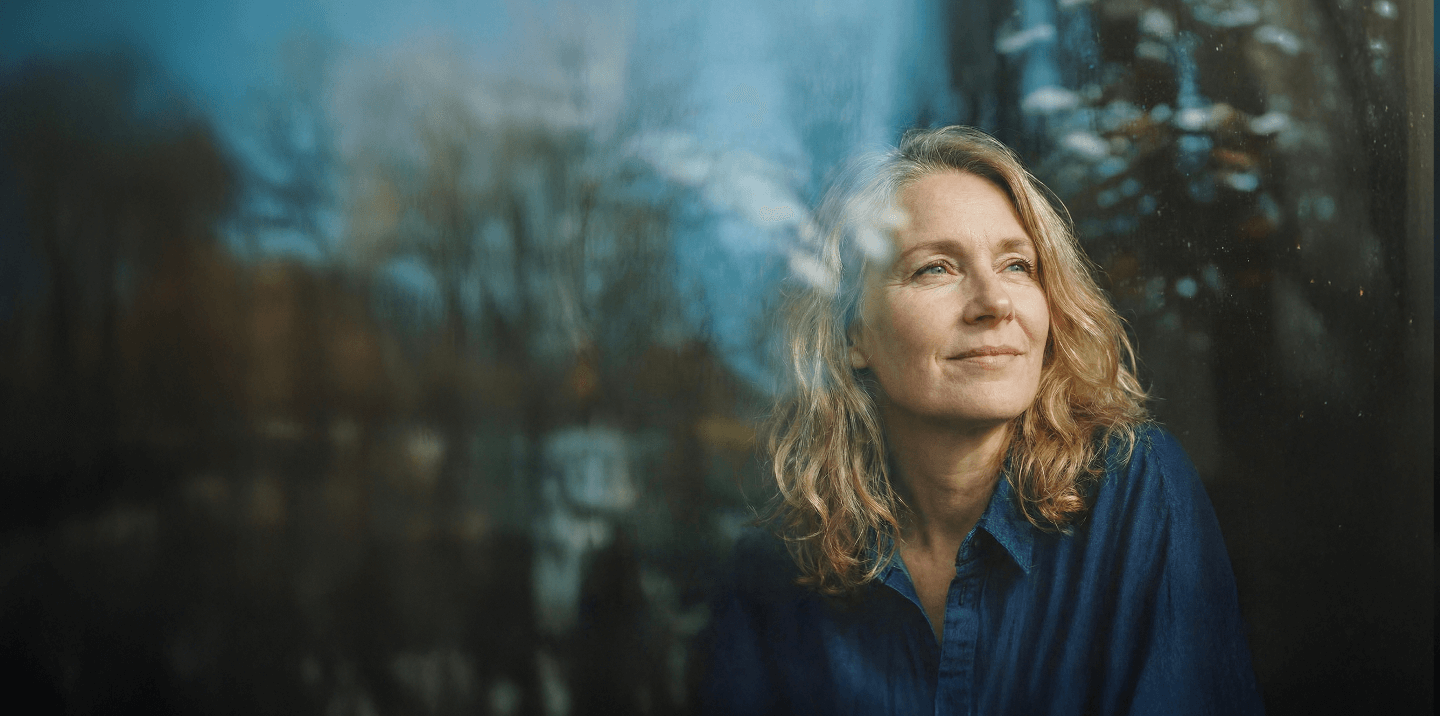 Woman with blonde hair gazing happy and thoughtfully out a window, with soft reflections and blurred trees outside